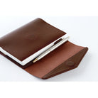 A brown leather notebook cover holding a white notebook and a silver pen, photographed on a clean white background.