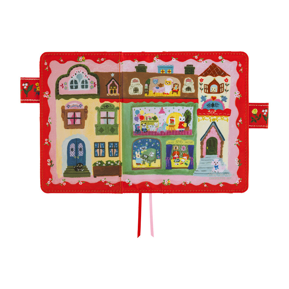 A children's fabric book or activity book featuring a colorful embroidered village scene on the cover, with a red border and ribbon tabs hanging from the bottom center, shown on a plain white background.