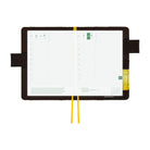 An open weekly planner with grid pages, held by a yellow elastic loop, set against a solid white background.
