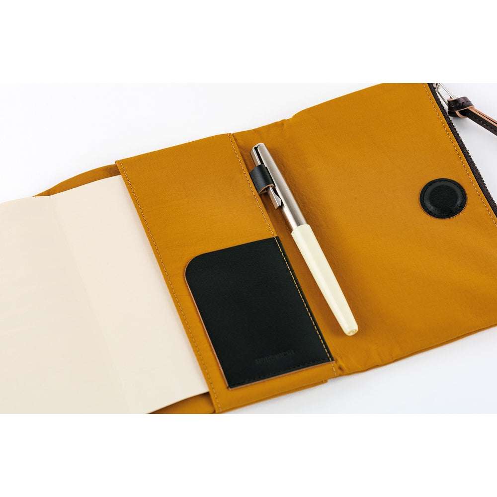 A mustard yellow canvas and leather notebook pouch shown open on a white background, holding a cream pen and a black notebook insert.