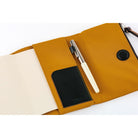 A mustard yellow canvas and leather notebook pouch shown open on a white background, holding a cream pen and a black notebook insert.