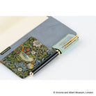 A black pen with gold accents resting in a floral-patterned pocket of a gray folio. The pocket features a detailed green and orange bird-and-floral print, and the pen clip is light green with gold lettering.