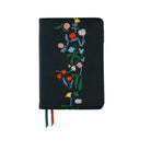 A black journal with colorful embroidered wildflowers on the front cover, featuring two ribbon bookmarks in green, white, and red hanging from the bottom edge, set against a plain white background.