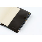 A close-up of a black leather notebook cover with a pen attached, shown open on a white background. The pen is black with gold accents, and the cover has a debossed logo near the bottom.