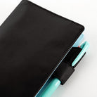 A close-up of a black fabric planner cover with a black elastic pen loop holding a mint green pen, on a white background.