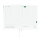 An open planner or notebook with a red cover and white grid pages, showing a weekly layout with date headers, tabs, and ribbon bookmarks on a plain white background.