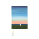 A hardcover notebook with a pastel landscape illustration on the cover, featuring a blue sky with a crescent moon, stars, a pink horizon, green field, and a small figure holding a string. The notebook is shown upright on a plain white background with a thin blue and cream ribbon bookmark hanging from the bottom.
