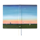 A hardcover notebook with a colorful landscape illustration on the cover, showing a blue sky with stars and a crescent moon over a green field at sunset. The notebook is open to show two pages with printed text, and has a blue ribbon bookmark hanging from the spine.