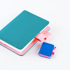 A teal notebook with a pink pen loops and a small matching blue-and-pink notebook-shaped accessory attached by a chain, photographed on a clean white background.