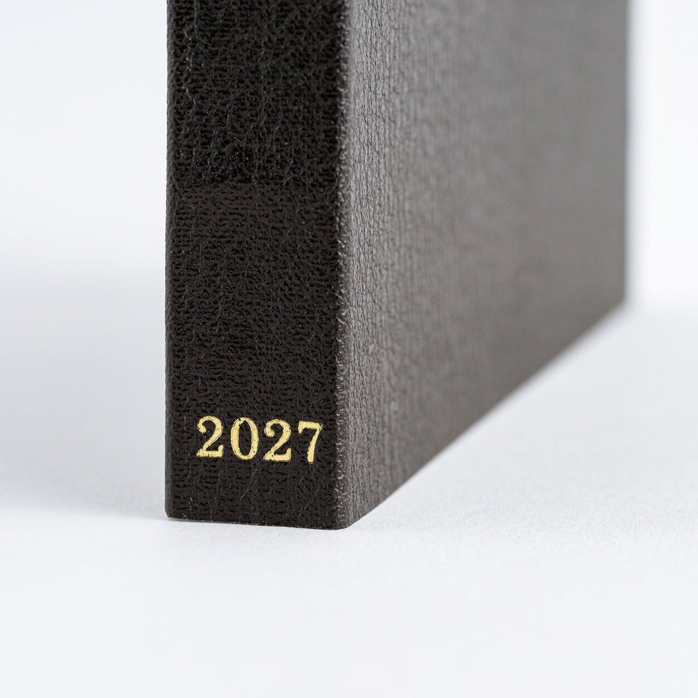 Close-up of a dark textured 2027 planner standing upright, showing the gold-foiled year '2027' on the spine against a clean white background.
