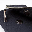 Close-up of a black fabric pouch with a silver zipper, a light tan leather pull tab, and two round black snap buttons on a white background.