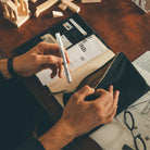 A close-up overhead view of a person's hands opening a black zippered pouch on a wooden desk, with papers and a pen visible inside and around it.