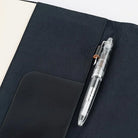 A close-up of a black fabric notebook cover with a clear pen resting inside a black pocket on a white surface.
