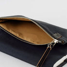 A close-up of a black zippered pouch shown open, revealing a tan interior lining and a gold-toned zipper. A small leather tag with a monogram logo is attached near the zipper pull.