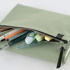 A sage green canvas zippered case shown open, filled with stationery items including pens, a correction tape dispenser, a ruler, and sticky notes, on a clean white background.