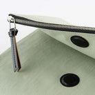 A close-up of a sage green fabric pouch with a black zipper, a black leather tassel zipper pull, and two black snap buttons.