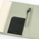 A sage green fabric notebook cover laid open on a white background, with a cream-colored notebook insert on the left, a black leather pocket on the lower left, and a silver pen resting on the right side.