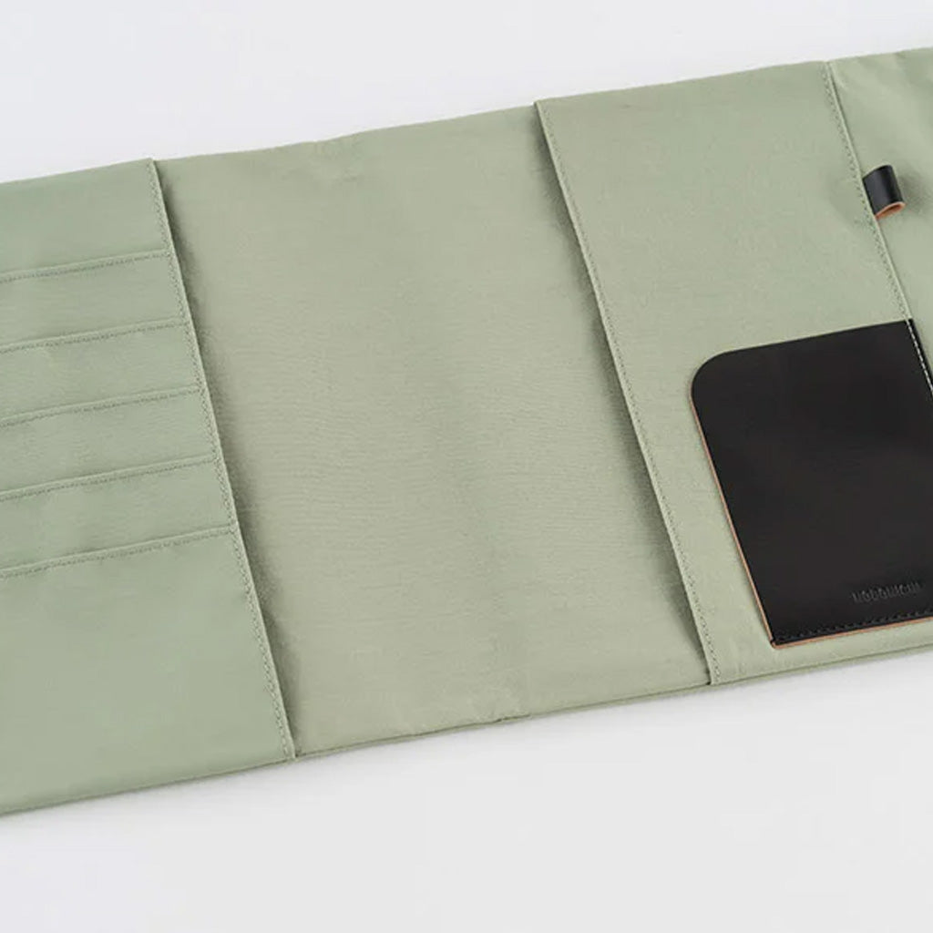 An open sage green fabric notebook cover with multiple compartments, a black leather card holder pocket, and a small black loop tab, photographed on a white background.