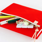 A bright red zippered pouch shown open on a white background, with pens, a ruler, and small office supplies peeking out. A small gold-toned logo tab is visible near the zipper pull.