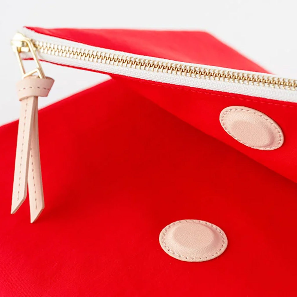 Close-up of a bright red zip pouch with a gold zipper, a light beige leather pull tab, and two oval light beige patches on a white background.