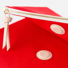 Close-up of a bright red zip pouch with a gold zipper, a light beige leather pull tab, and two oval light beige patches on a white background.