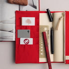 A red fabric organizer pouch laid open on a light gray surface, holding cards, a ruler, a pen, and a notebook page. The pouch has multiple pockets and a zipper closure.