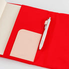 A red fabric notebook cover shown open on a white background, with a cream-colored interior page, a pale blush pocket, and a white pen tucked inside.