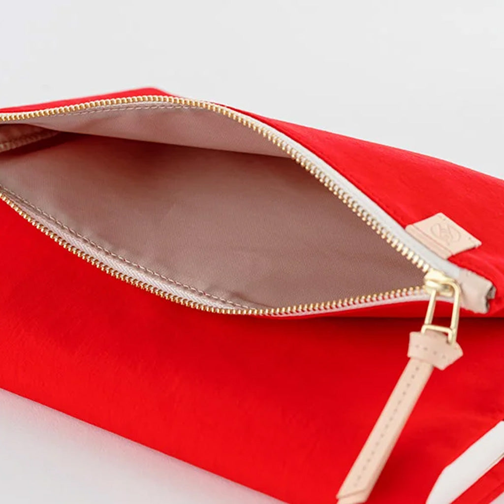 A close-up of a bright red zippered pouch shown open, revealing a light beige interior lining and a gold-tone zipper. A small beige leather pull tab is visible on the right side against a clean white background.