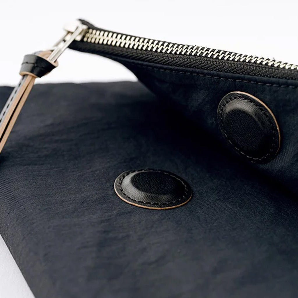 Close-up of a black fabric pouch with a silver zipper, black leather trim, and two circular black leather patches on a white background.