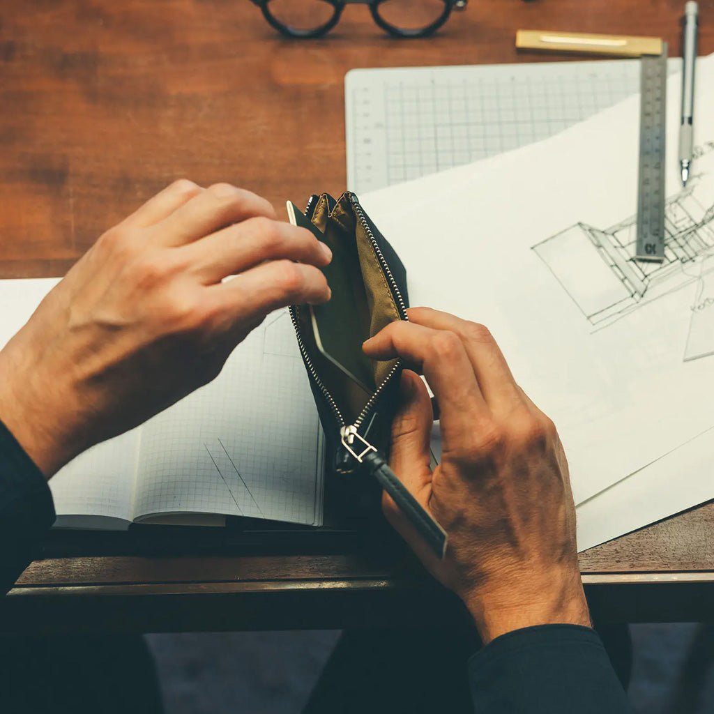 A close-up of a person's hands opening a black leather zippered pouch and inserting a notebook, set against a desk with architectural drawings, a ruler, and glasses.