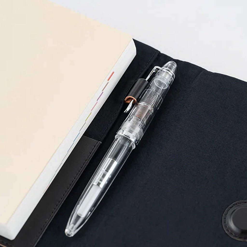A transparent pen with a black clip and internal ink cartridge, resting on a black fabric surface next to a cream-colored notebook.