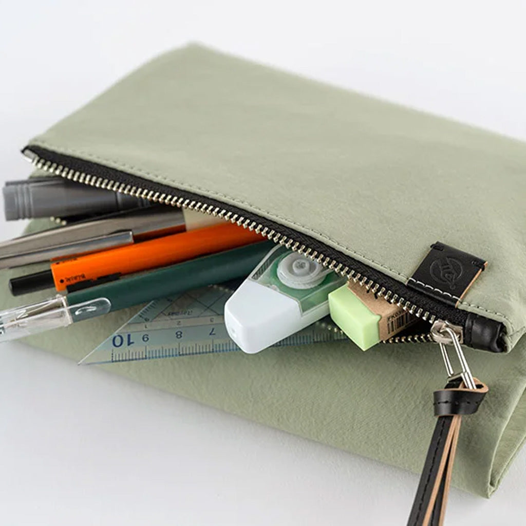 An olive green fabric zippered pouch with a black zipper and a small black brand tag, shown open with pens, a correction tape dispenser, a ruler, and a eraser inside on a white background.