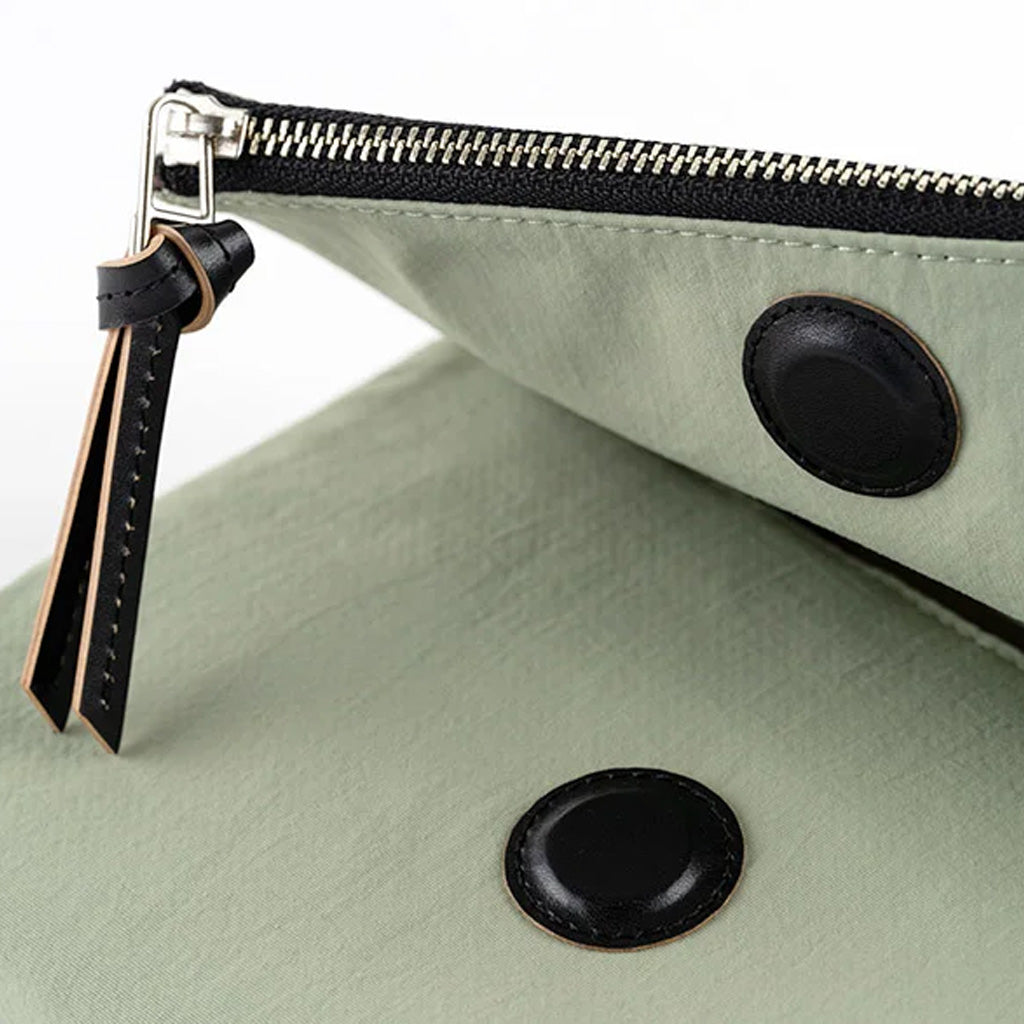 A close-up of a sage green pouch with black leather trim, a black zipper, and two black circular snap details, shown on a white background.