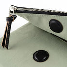 A close-up of a sage green pouch with black leather trim, a black zipper, and two black circular snap details, shown on a white background.