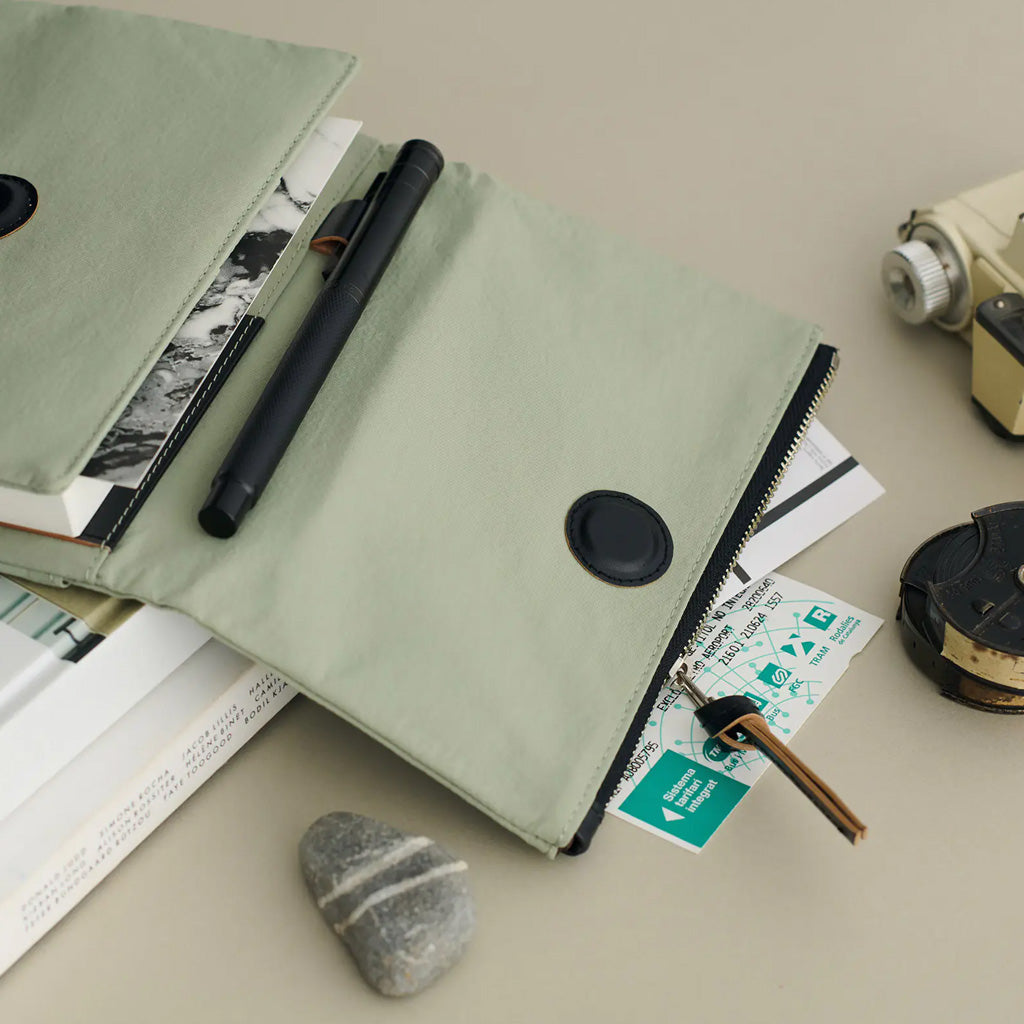 A sage green zippered notebook cover with a black snap button, shown open and styled with a black pen, books, a key, and a small stone on a neutral beige surface.
