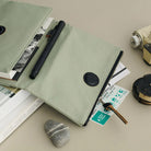 A sage green zippered notebook cover with a black snap button, shown open and styled with a black pen, books, a key, and a small stone on a neutral beige surface.