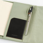 An open sage green notebook cover with a black leather pocket and a silver pen, photographed on a clean white background.
