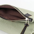 Close-up of a sage green leather pouch with an open zipper revealing a dark brown interior, featuring a silver-tone metal zipper pull and a small black leather brand tab.