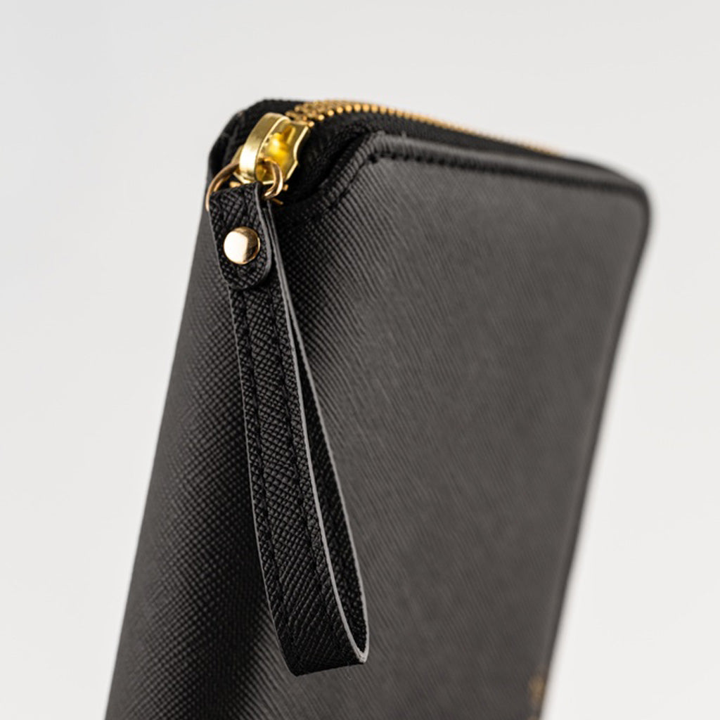 Close-up of a black textured zip-around notebook cover with a gold-tone zipper pull and wrist strap on a light gray background.