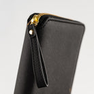 Close-up of a black textured zip-around notebook cover with a gold-tone zipper pull and wrist strap on a light gray background.