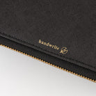 Close-up of a black textured fabric pouch with a gold zipper and gold 'handwrite' logo printed on the side, set against a light gray background.