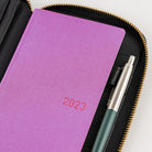 A close-up of a purple textured 2023 planner or notebook inside a black zippered case with gold-tone zipper, with a pen tucked in beside it.
