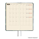 An open planner showing a monthly calendar spread with cream grid pages. A ribbon bookmark hangs from the bottom.