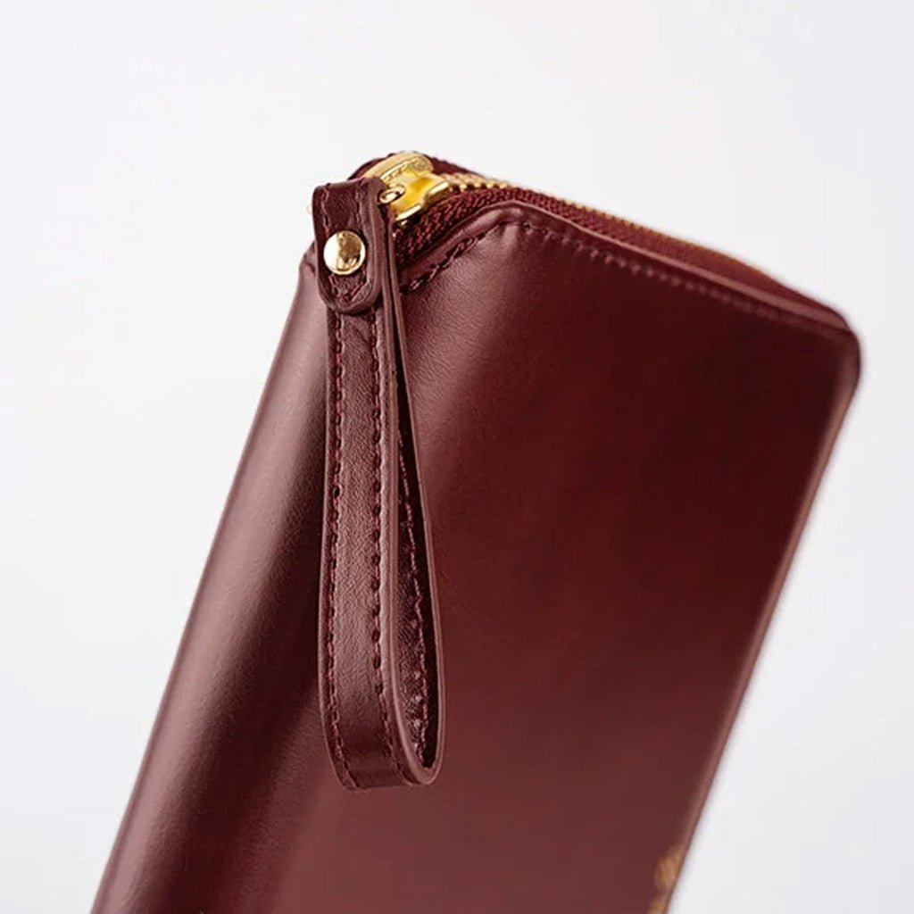 A close-up of a dark burgundy leather notebook zip case with a wrist strap and gold-tone zipper hardware, shown on a clean white background.