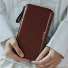 A person in a white robe holds a dark brown leather zip-around notebook case with a wrist strap and gold logo detailing.