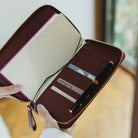 A brown zip-around Weeks planner case is shown open and held by hands, displaying its interior card slots, pen pocket, and lined paper compartments. The wallet has a gold zipper, a purple interior trim, and is photographed in a warm indoor setting.