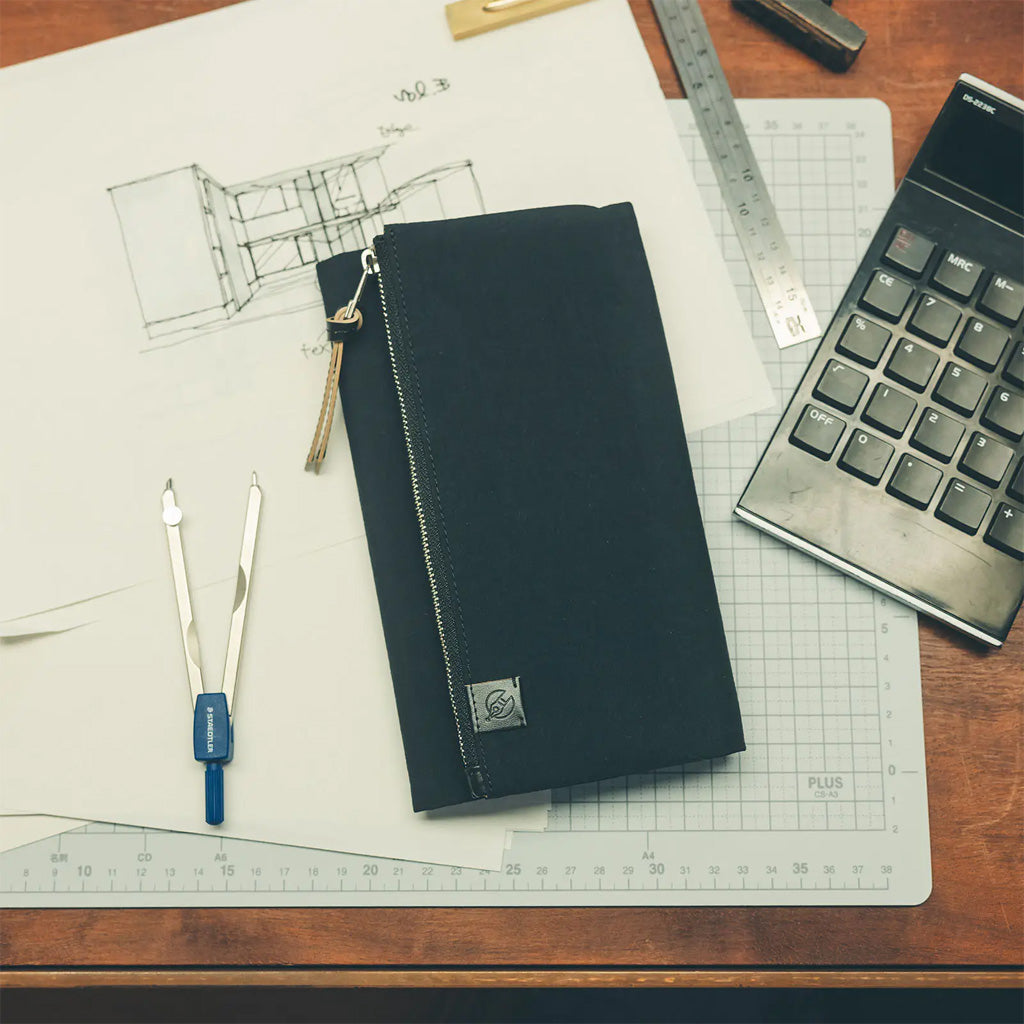 A black zippered fabric notebook cover with a small logo patch, placed on a desk with drafting tools, a calculator, and a grid cutting mat.