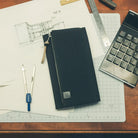 A black zippered fabric notebook cover with a small logo patch, placed on a desk with drafting tools, a calculator, and a grid cutting mat.