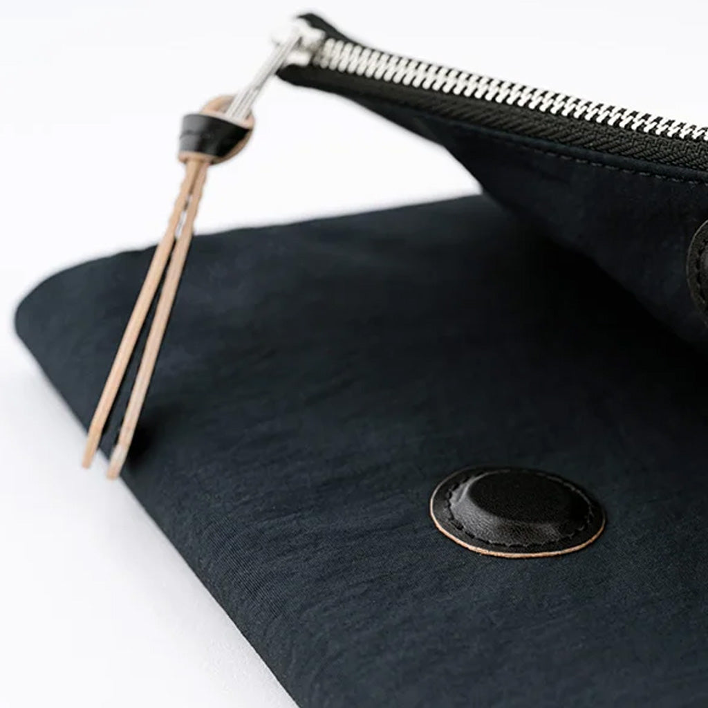 Close-up of a black fabric pouch with a silver zipper, a black leather pull tab, and a round black button closure on a white background.