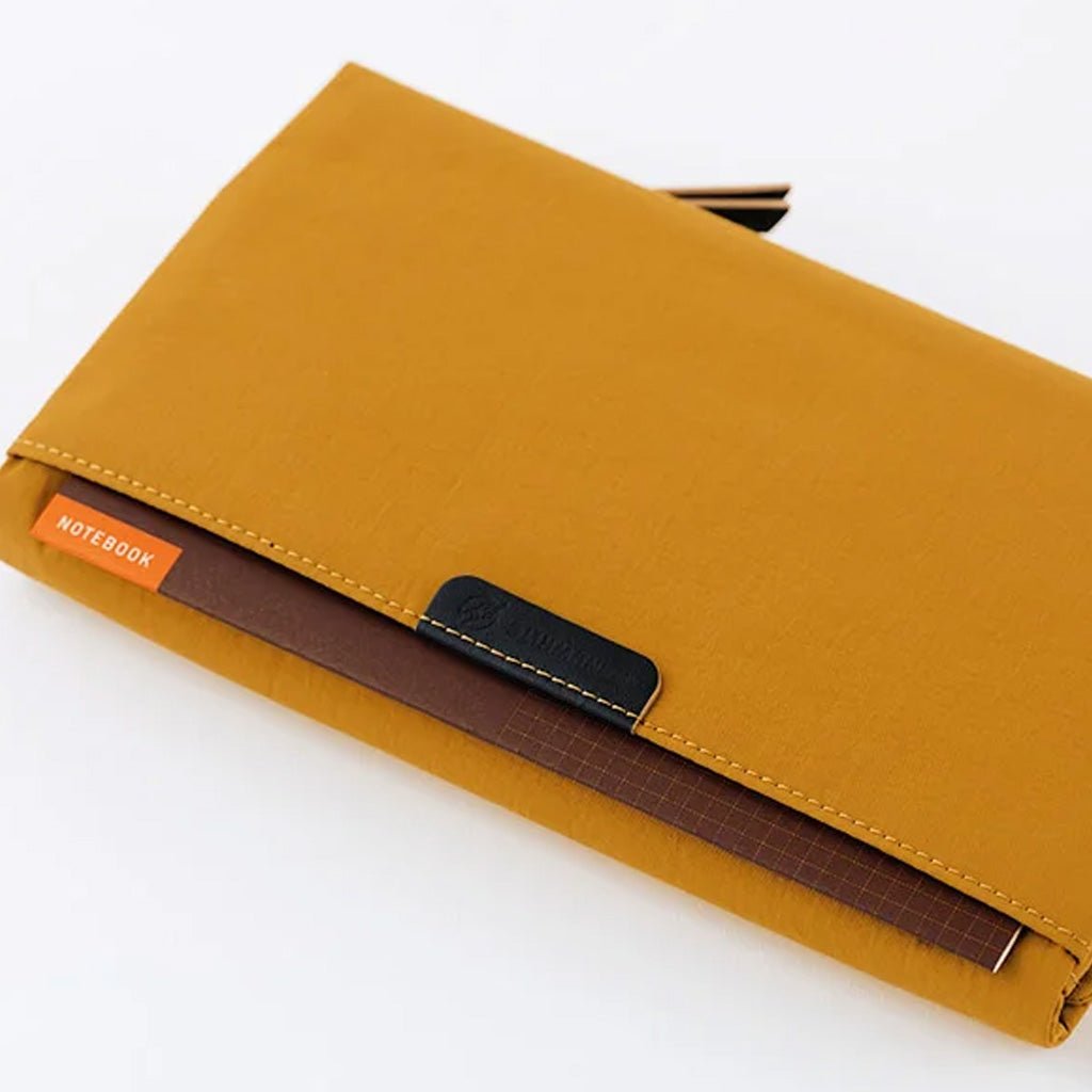 A mustard yellow notebook cover with a black leather brand tab and an orange NOTEBOOK inside pocket, shown on a clean white background.